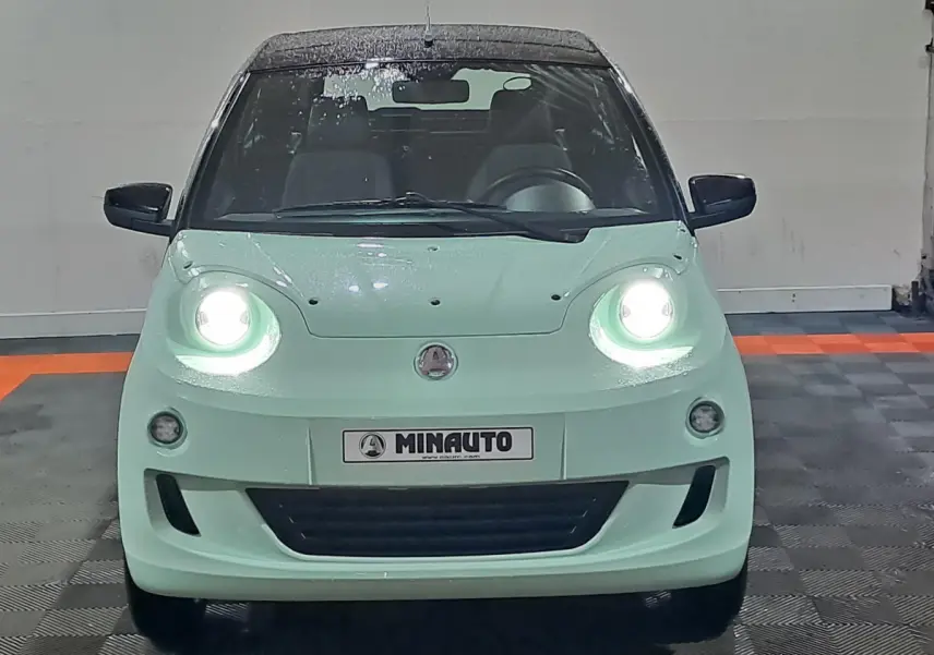Vue frontale d'une Aixam MINAUTO E chic 2026 verte avec phares allumés et rétroviseurs noirs à caméra côté conducteur.