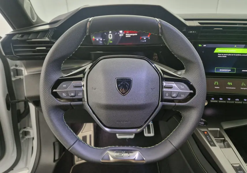 Vue rapprochée du volant cuir perforé noir avec logo Peugeot et badge GT de la 308 blanche 2025, tableau de bord digital visible.