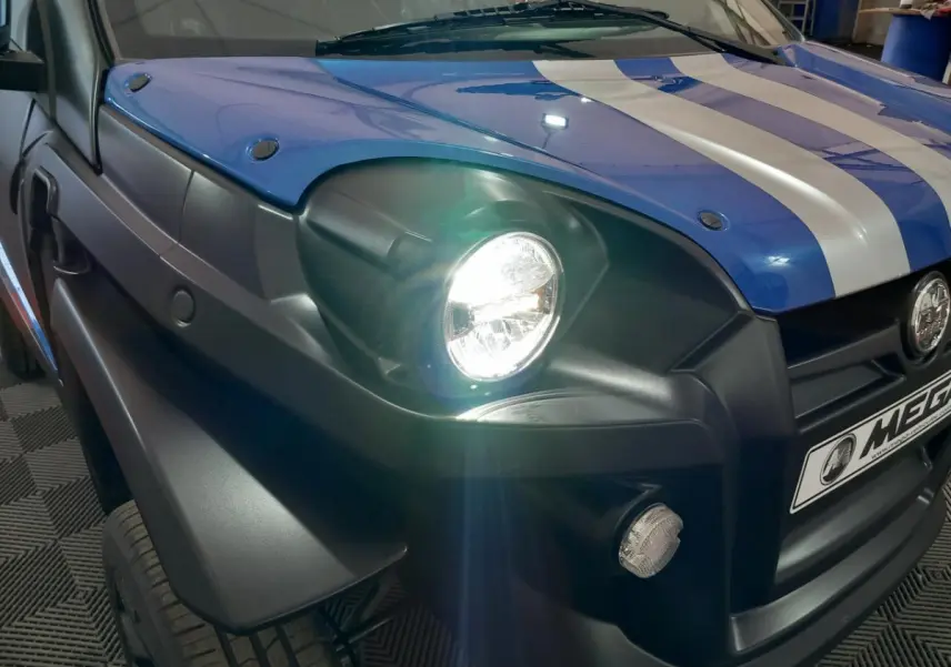 Gros plan sur l’avant gauche bleu électrique de l’AIXAM MEGA E-SCOUTY EVO avec phare allumé et bandes blanches sur le capot.