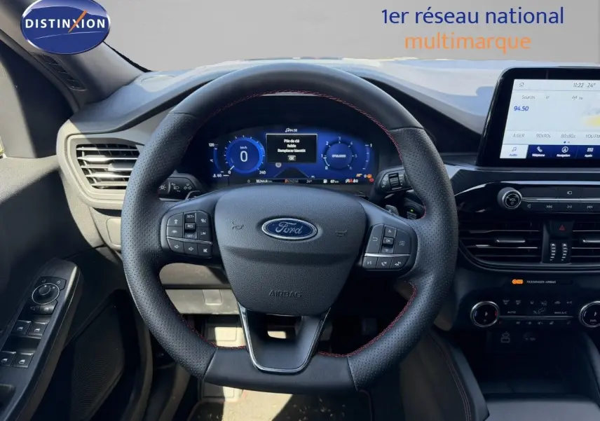 Vue intérieure centrée sur le volant noir perforé avec surpiqûres rouges du Ford Kuga 2024, tableau de bord digital et écran tactile.