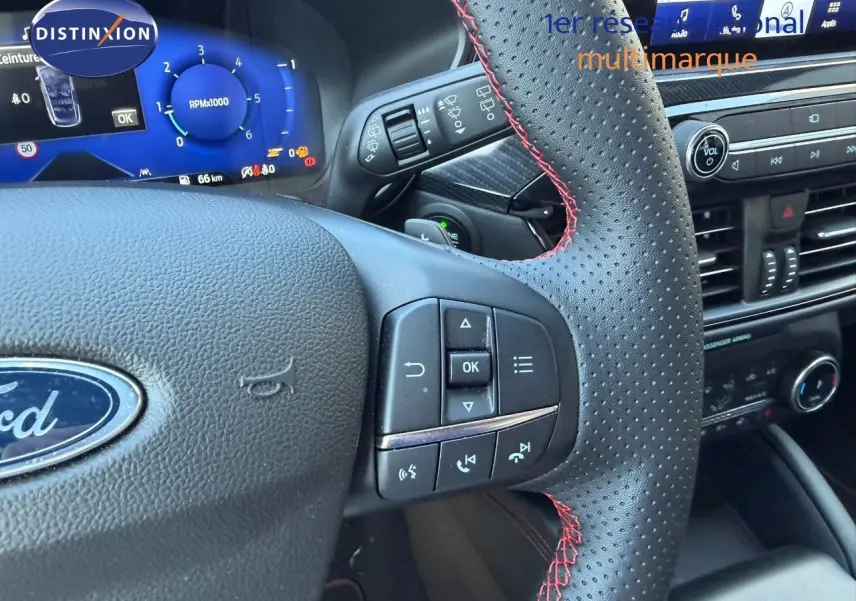 Gros plan sur le volant en cuir perforé avec surpiqûres rouges du Ford Kuga 2024, tableau de bord digital bleu en arrière-plan.