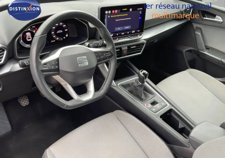 Vue intérieure du poste de conduite de la SEAT LEON 2025, volant cuir multifonction et levier de vitesse manuel, sièges tissu gris clair.