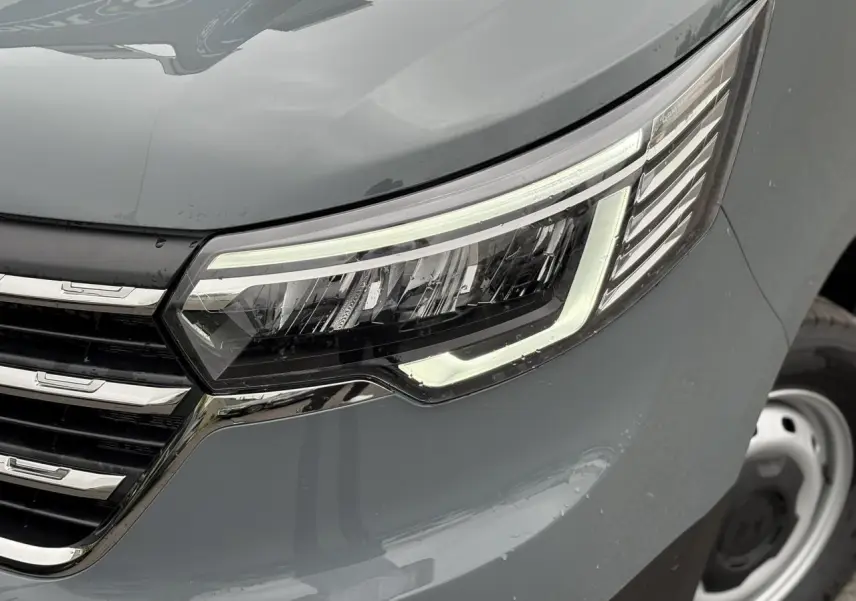 Gros plan sur le phare avant droit du Renault Trafic Fourgon gris urbain, soulignant la calandre chromée et la roue acier.
