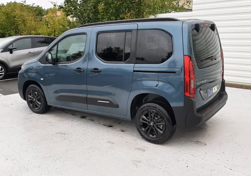 Vue 3/4 arrière droite du Citroën Berlingo bleu Kiama avec vitres teintées et jantes noires sur fond urbain.