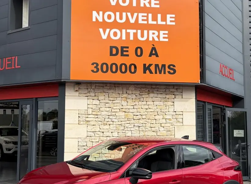 Renault Clio Evolution rouge vue en 3/4 avant droit, avec jantes noires et calandre distinctive, devant un bâtiment commercial.