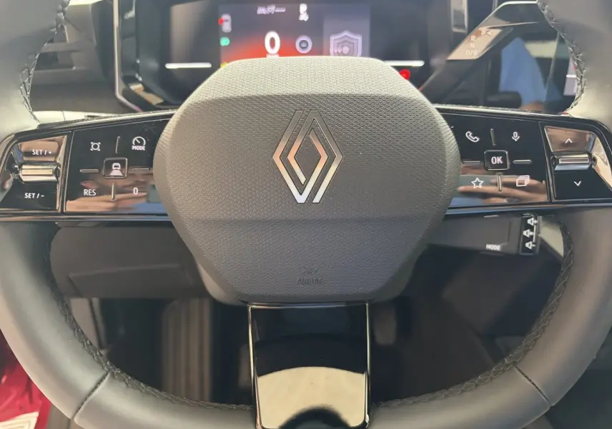 Gros plan sur le volant multifonction noir avec logo Renault, tableau de bord numérique flou en arrière-plan.