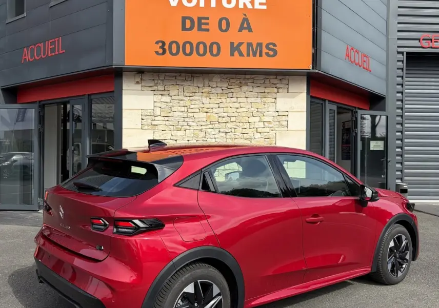 Vue 3/4 arrière droite d'une Renault Clio Evolution rouge 2025 avec toit noir et jantes noires, garée devant un bâtiment commercial.