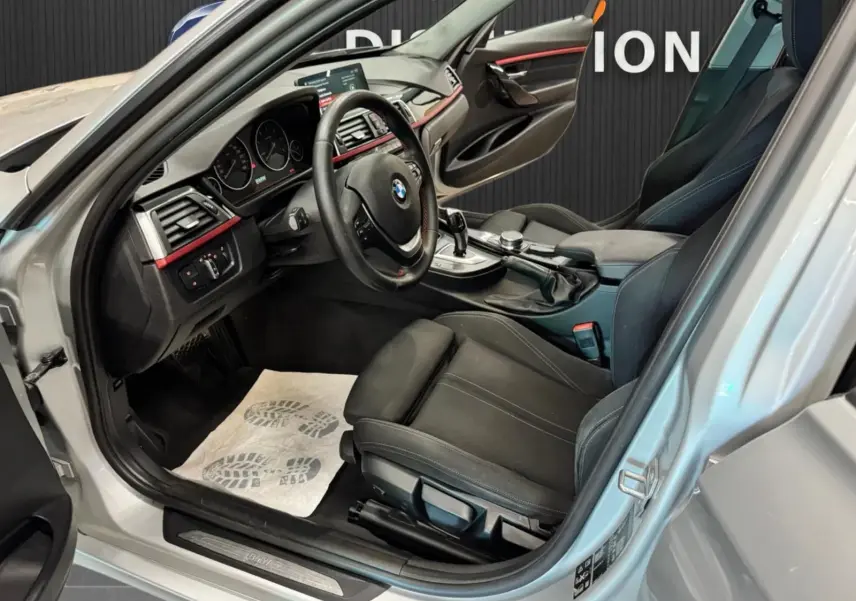 Vue intérieure côté conducteur d'une BMW Série 3 Touring gris aluminium, avec sièges noirs et tableau de bord sportif.