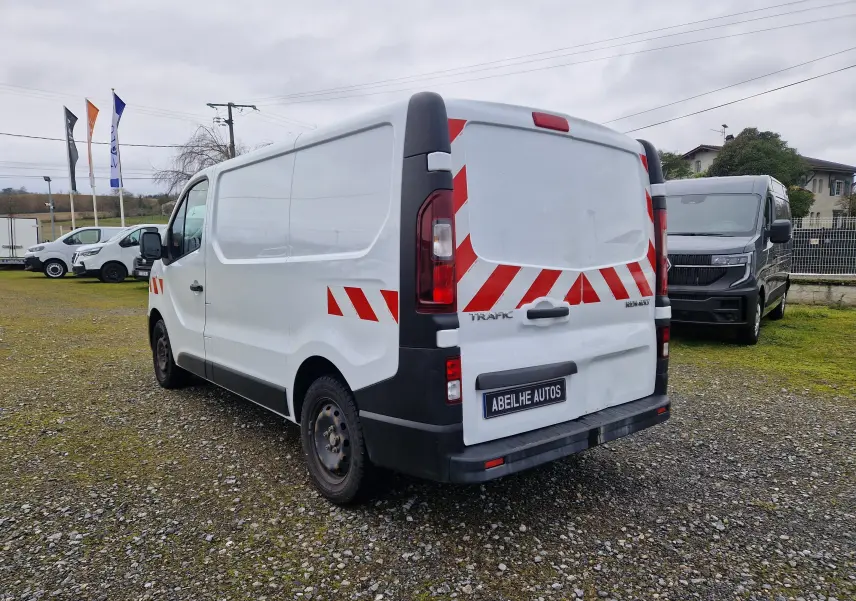 Vue 3/4 arrière droite d’un Renault Trafic blanc avec bandes rouges réfléchissantes sur un terrain gravillonné.