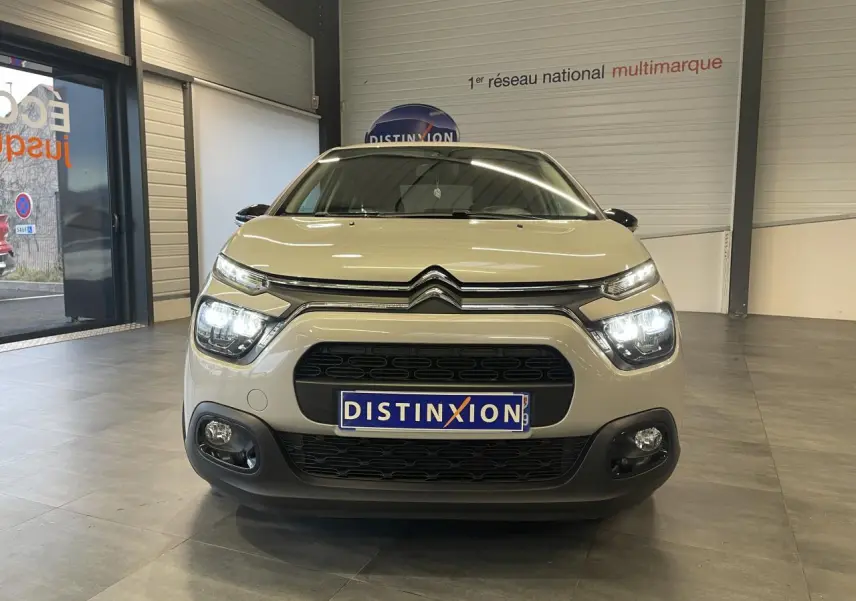 Vue frontale d'une Citroën C3 beige clair 2024 avec feux allumés et calandre noire en showroom.