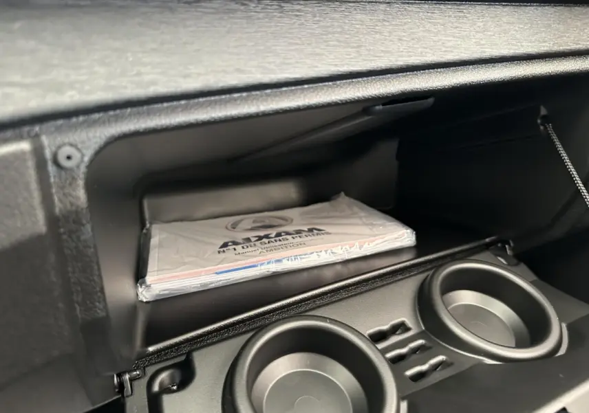 Gros plan sur la boîte à gants ouverte de l'AIXAM AMBITION 0.5 CITY PACK 2026 avec manuel visible et porte-gobelets intégrés.