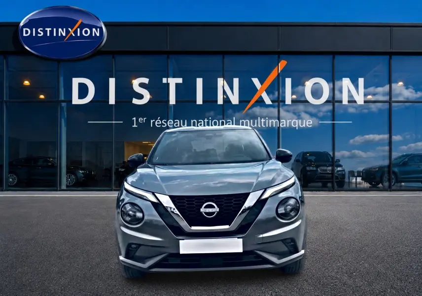 Nissan Juke gris 2025 en vue frontale, avec calandre noire et phares ronds distinctifs, devant showroom Distinxion.