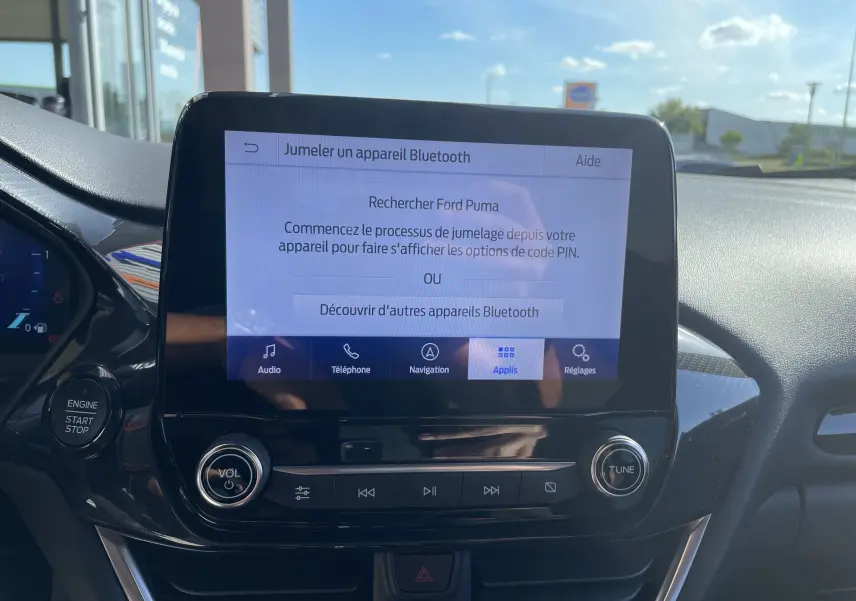 Écran tactile central du Ford Puma 2021 affichant le jumelage Bluetooth, avec boutons de volume et réglage sous un ciel ensoleillé.