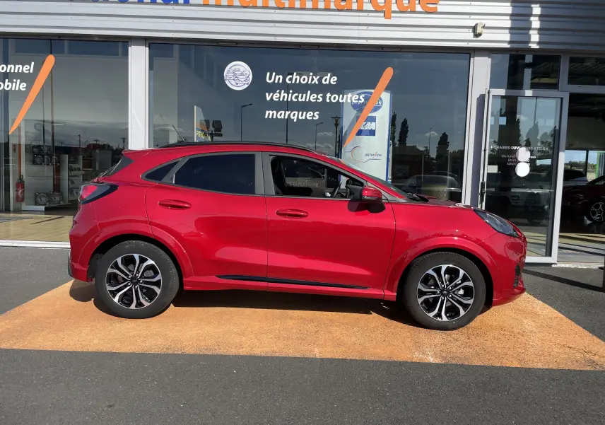 Vue latérale droite d'un Ford Puma rouge 2021 avec jantes bi-ton et vitres teintées, garé devant un showroom.