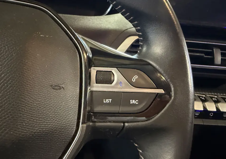 Gros plan sur les commandes gauche du volant noir d’un Peugeot 5008 gris, avec boutons LIST, SRC et téléphone.