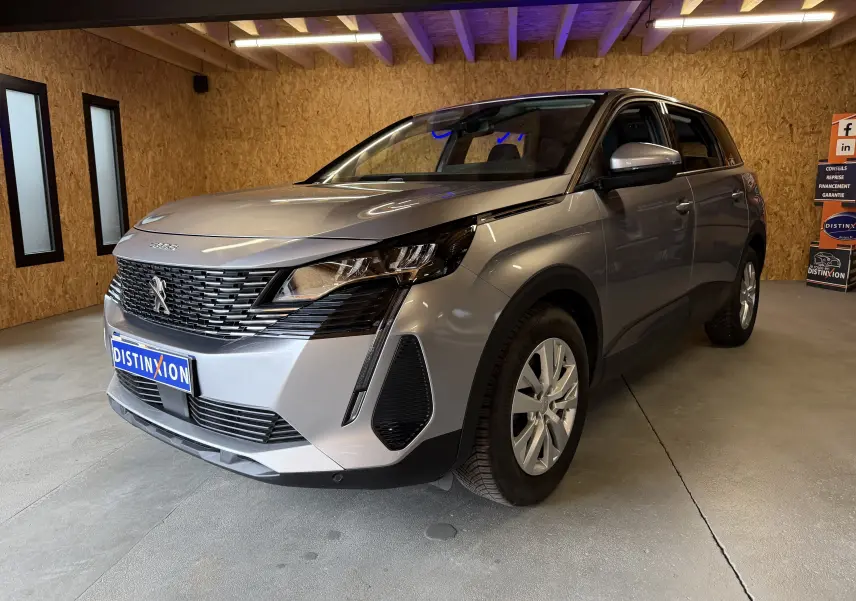 Peugeot 5008 gris en 3/4 avant droit dans un intérieur bois, mettant en valeur sa calandre noire et ses phares modernes.