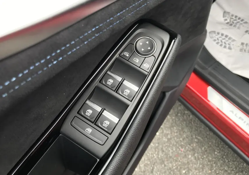 Gros plan sur la commande des vitres côté conducteur avec surpiqûres bleues et seuil de porte rouge Alpine visible.