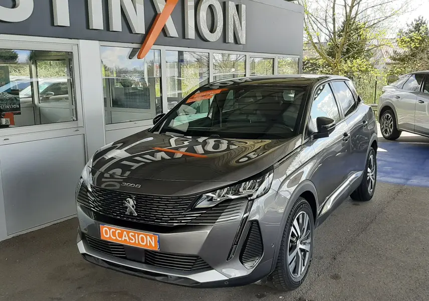 Peugeot 3008 gris platinium en 3/4 avant droit, avec calandre noire et feux LED distinctifs, stationné devant un mur blanc.