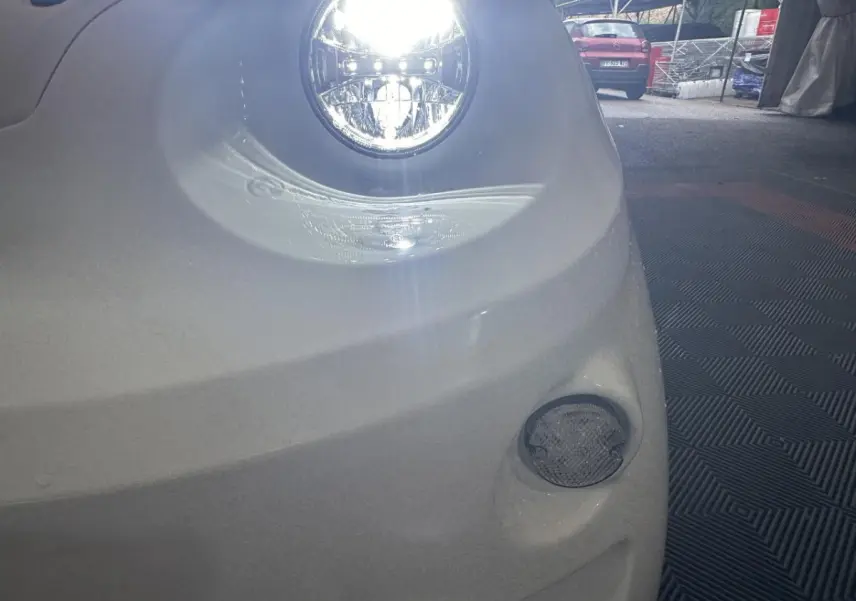 Gros plan sur le phare avant droit allumé d'une Aixam Minauto E Access blanche, avec un feu antibrouillard visible en bas.