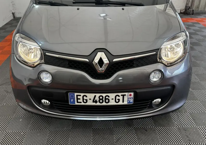 Vue frontale rapprochée d'une Renault Twingo gris foncé avec phares allumés et logo chromé bien visible.