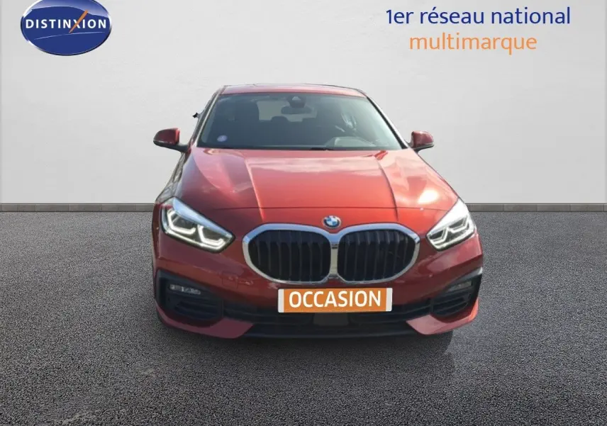 Vue avant d'une BMW Série 1 116i orange Sunset Metal avec phares LED allumés sur fond neutre.