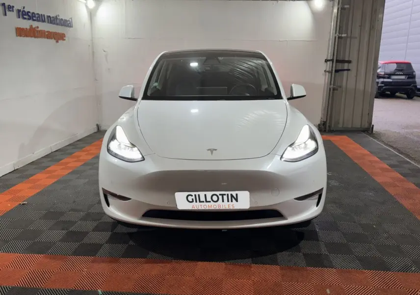 Vue avant d'une Tesla Model Y blanche avec phares allumés dans un garage éclairé.
