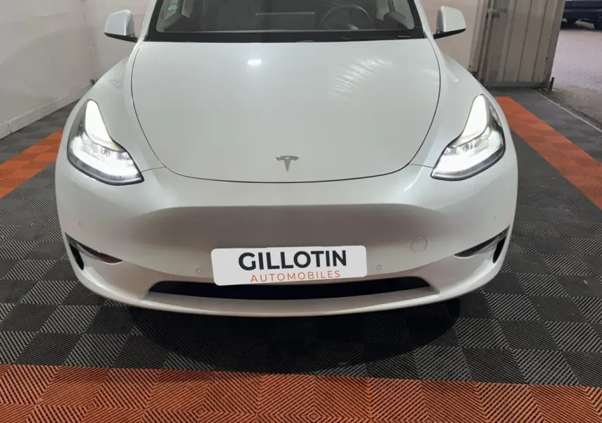 Vue avant d'une Tesla Model Y blanche avec phares LED allumés, sur sol damier noir et orange en intérieur.