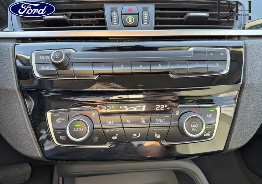 Tableau de bord noir brillant d'une BMW X1 sDrive18dA 2018, vue rapprochée sur la console de climatisation et audio.