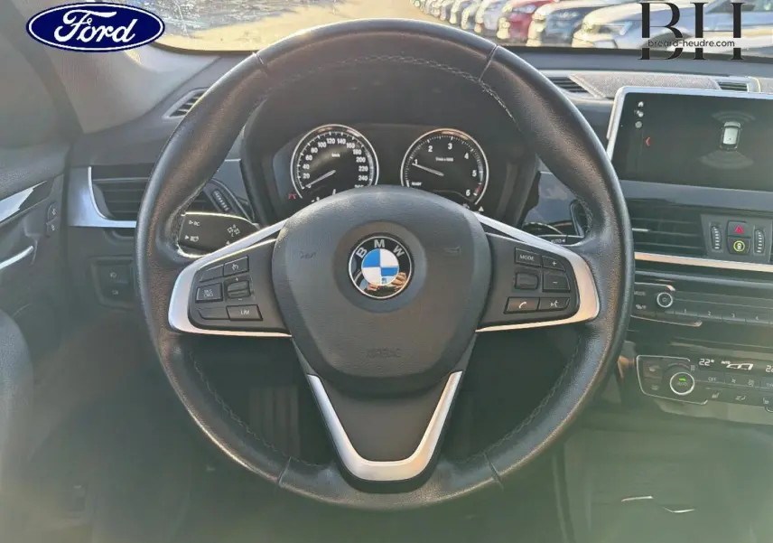 Vue intérieure centrée sur le volant cuir multifonction d'une BMW X1 noire avec tableau de bord et écran multifonction.