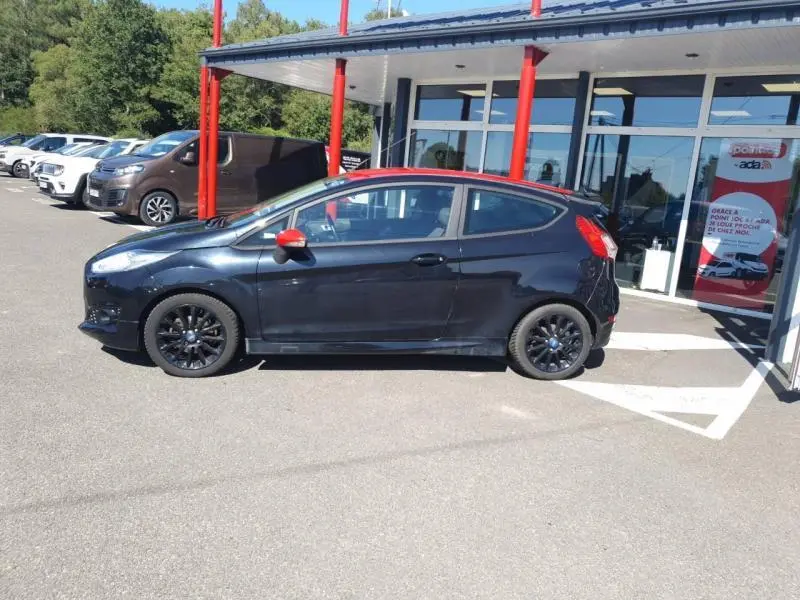 Ford Fiesta noire vue de profil côté gauche, avec rétroviseur rouge et jantes noires sport devant un bâtiment moderne.