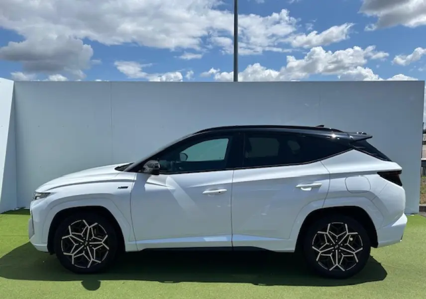 Profil côté gauche du Hyundai Tucson blanc polaire avec toit noir et jantes au design géométrique sous un ciel bleu.