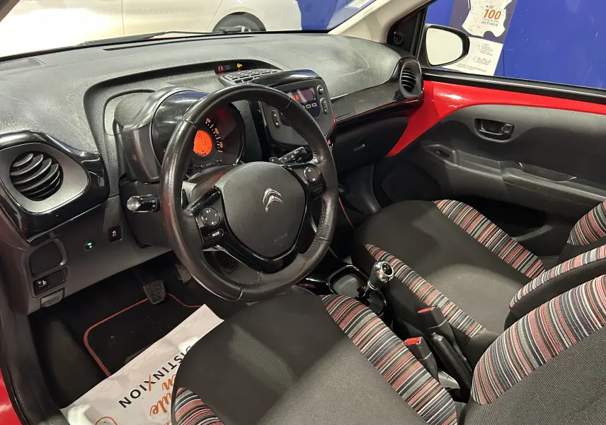 Vue intérieure côté conducteur de la Citroën C1 rouge 2019, volant multifonction et sièges tissu rayés rouges et noirs.