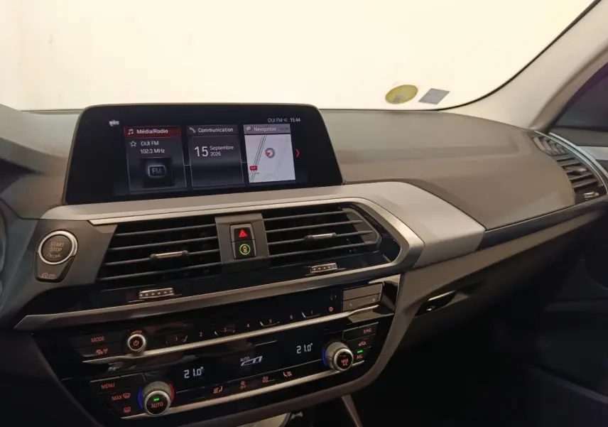 Tableau de bord intérieur de BMW X3 gris clair, vue de face mettant en valeur l’écran tactile et les commandes climatisation.