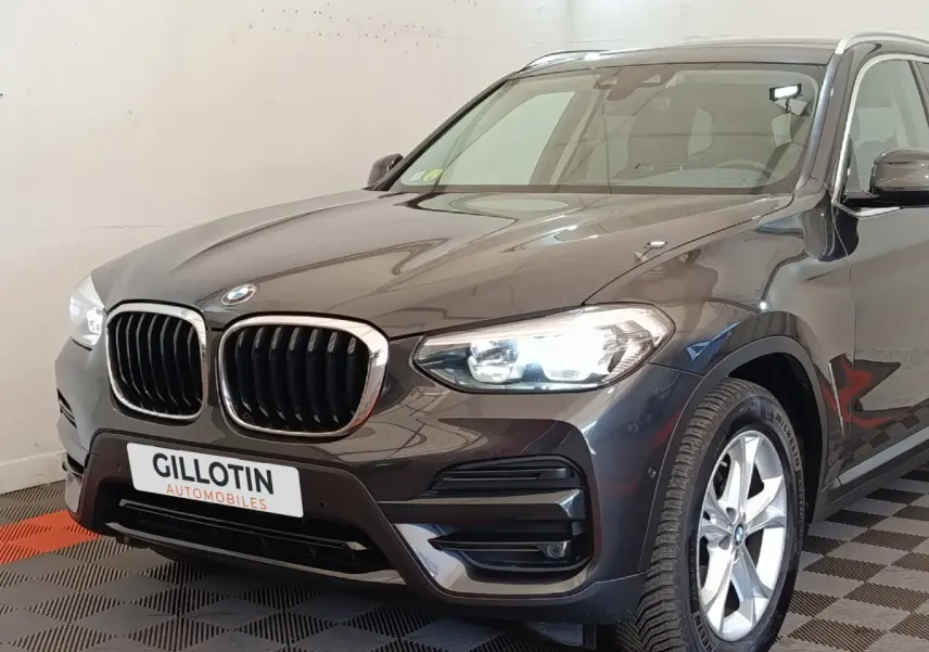 Vue 3/4 avant droite d’un BMW X3 gris clair avec calandre double haricot et phares allumés en intérieur showroom.