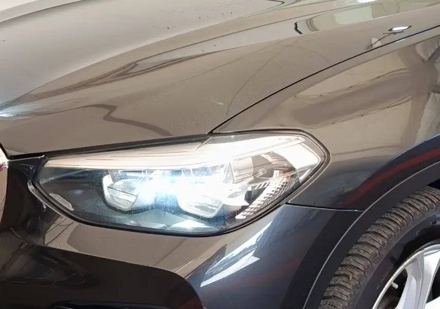 Gros plan sur le phare avant droit allumé d’un BMW X3 gris clair avec jante alu visible.