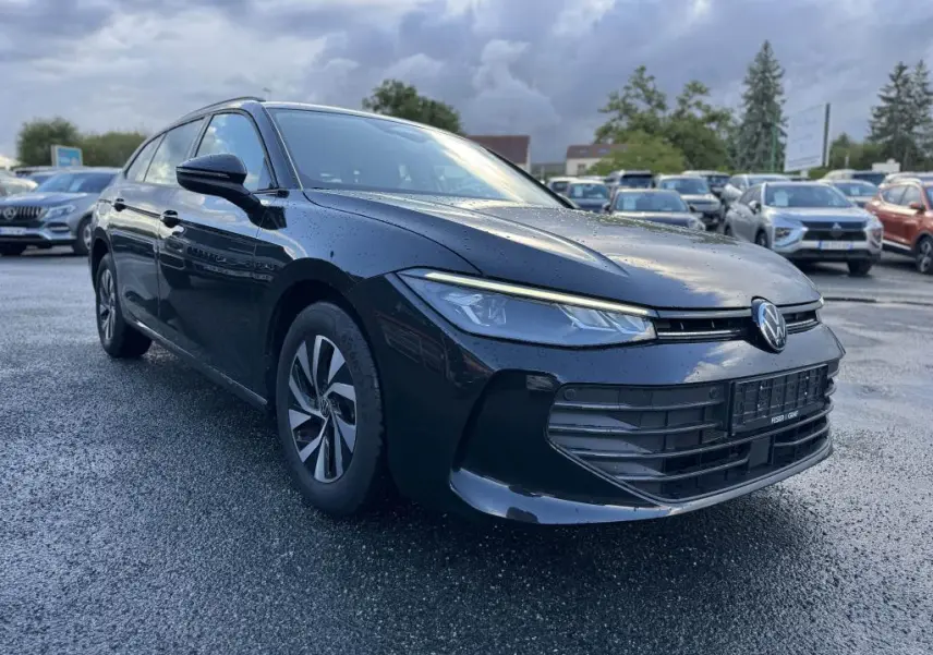 Volkswagen Passat SW noire vue en 3/4 avant gauche, avec phares LED et jantes alliage foncé sur parking humide.