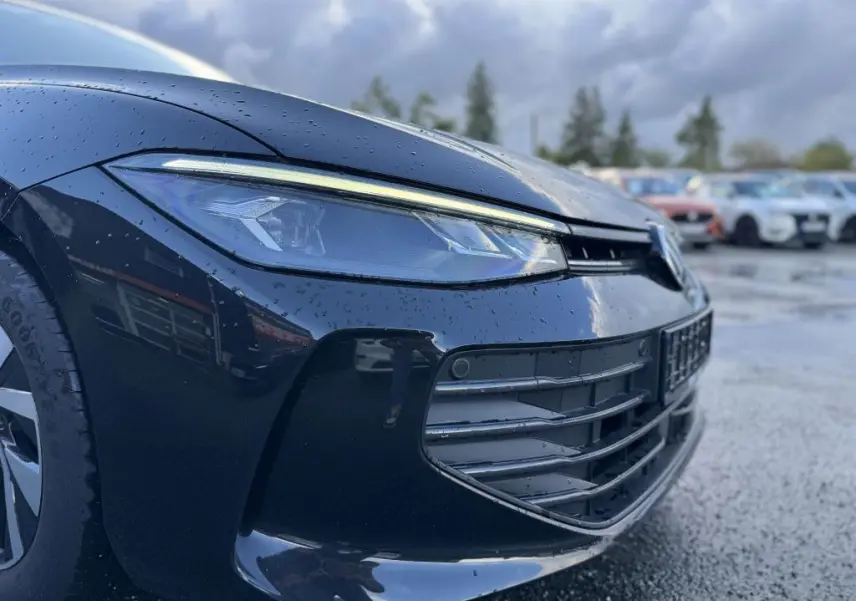 Gros plan sur le phare avant gauche d'une Volkswagen Passat SW noire 2024 avec gouttes de pluie et calandre visible.