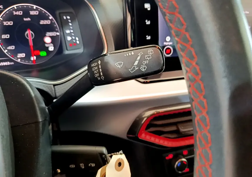 Gros plan sur la commande d’essuie-glace à gauche du volant cuir noir avec surpiqûres rouges, tableau de bord digital visible.