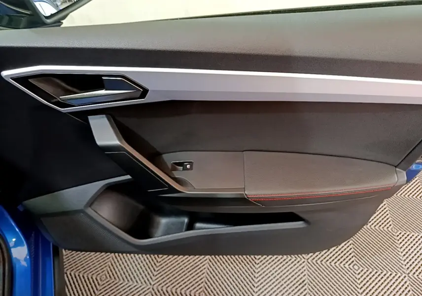 Intérieur côté gauche de la porte avant d’une SEAT Arona bleu 2022 avec poignée argentée et surpiqûres rouges.