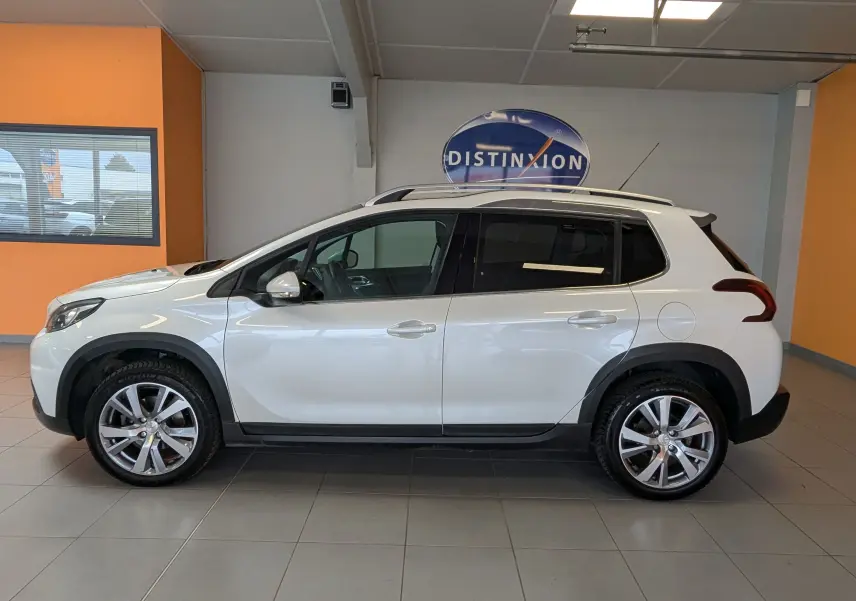 Vue de profil côté gauche d'un Peugeot 2008 blanc de 2017 avec jantes alliage et barres de toit en intérieur showroom.