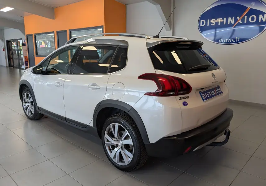 Vue 3/4 arrière droite d’un Peugeot 2008 blanc 2017 avec barres de toit et attelage dans un showroom.