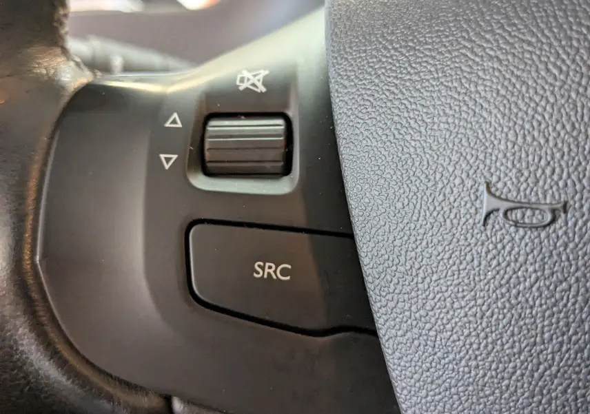 Gros plan sur les commandes noires du volant de la Peugeot 2008 blanche, bouton SRC et molette de volume visibles.