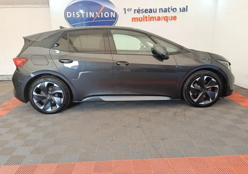 Vue de profil côté gauche d'une CUPRA BORN 230ch gris foncé avec jantes noires et toit spoiler en intérieur showroom.