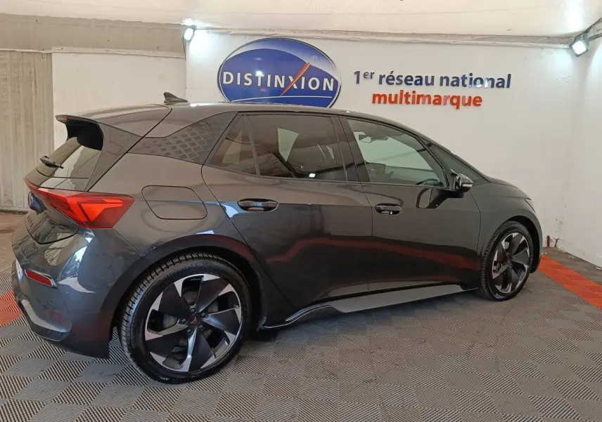 Vue latérale droite d'une CUPRA BORN 230ch gris foncé avec jantes noires et toit avec spoiler en intérieur showroom.
