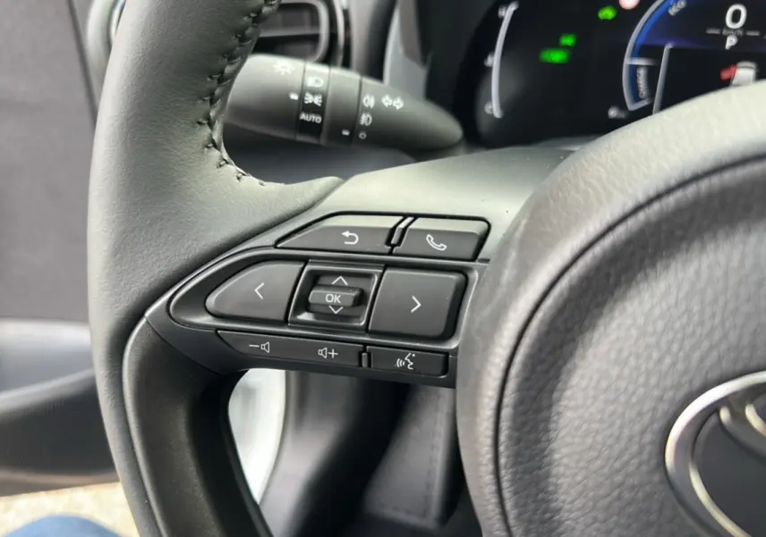 Gros plan sur les commandes du volant noir de la Toyota Yaris 1.5 Hybrid 116 BVA Design Plus 2025, tableau de bord en arrière-plan.