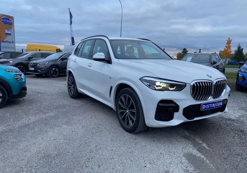 BMW X5 xDrive 30d blanc en 3/4 avant droit avec calandre M Sport et phares allumés sur parking extérieur.