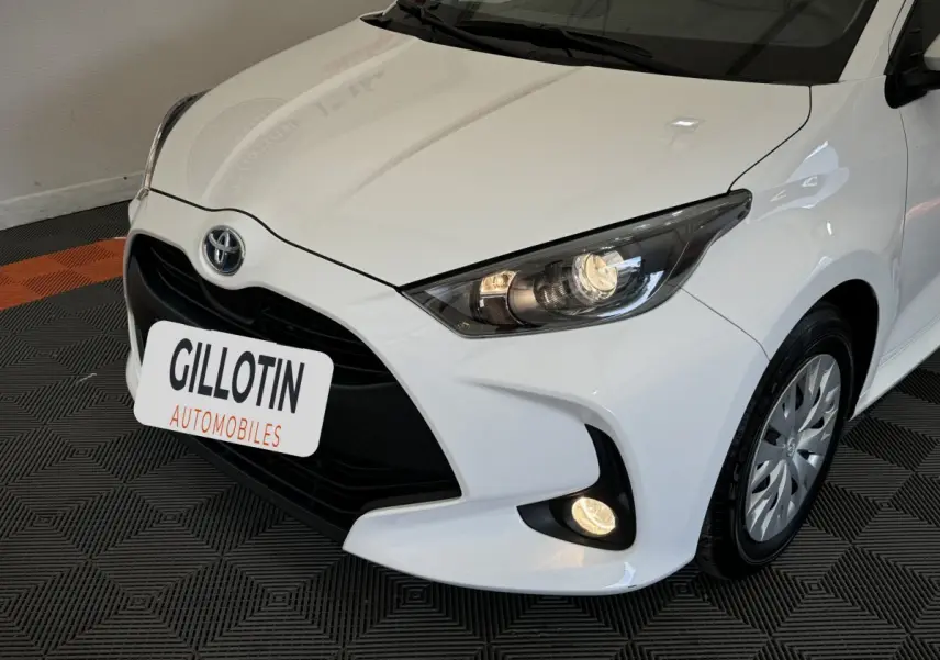 Vue 3/4 avant droite d'une Toyota Yaris blanche hybride 2022 avec phares allumés et calandre noire distinctive.