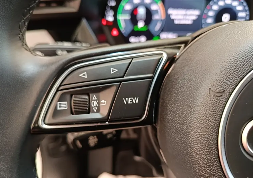 Gros plan sur les commandes gauche du volant cuir noir d’un Audi A3 hybride, avec tableau de bord numérique en arrière-plan flou.
