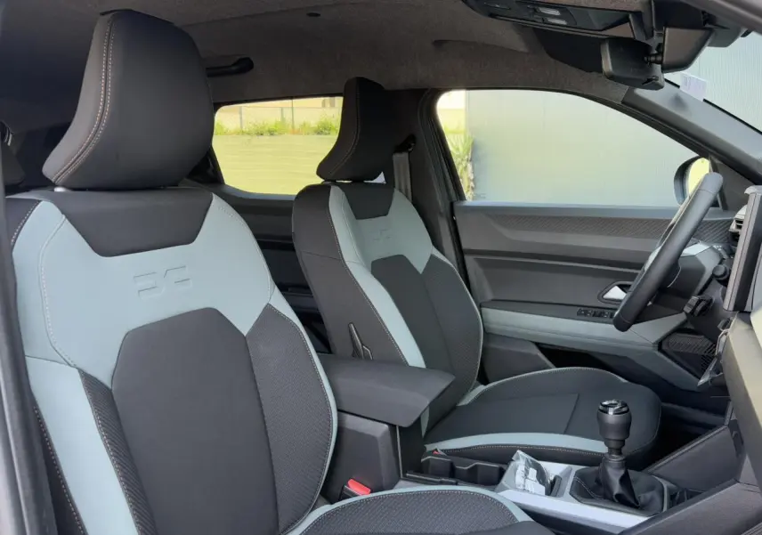 Vue intérieure côté conducteur du Dacia Duster 2025 avec sièges bi-ton gris clair et noir et levier de vitesse manuel.