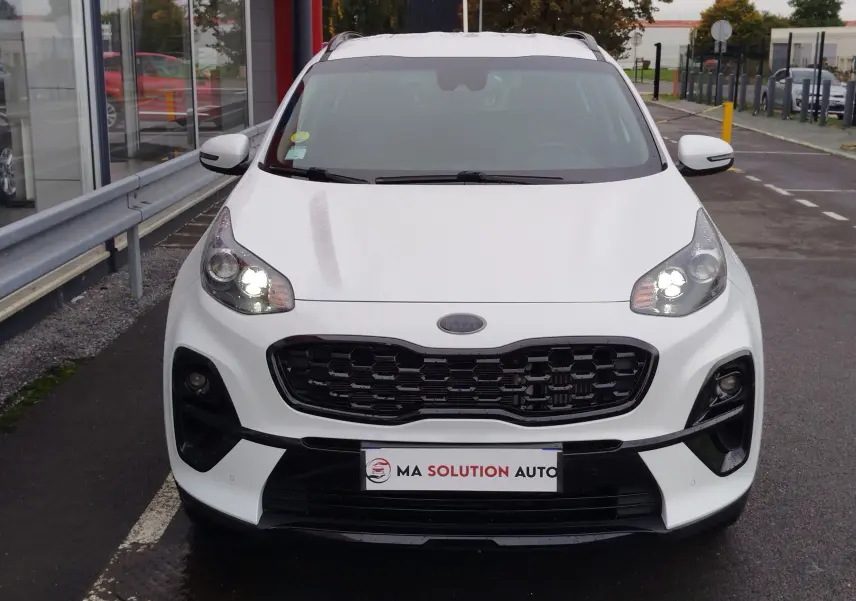 Vue frontale d'un KIA SPORTAGE blanc 2022 avec calandre noire et phares allumés sur parking.